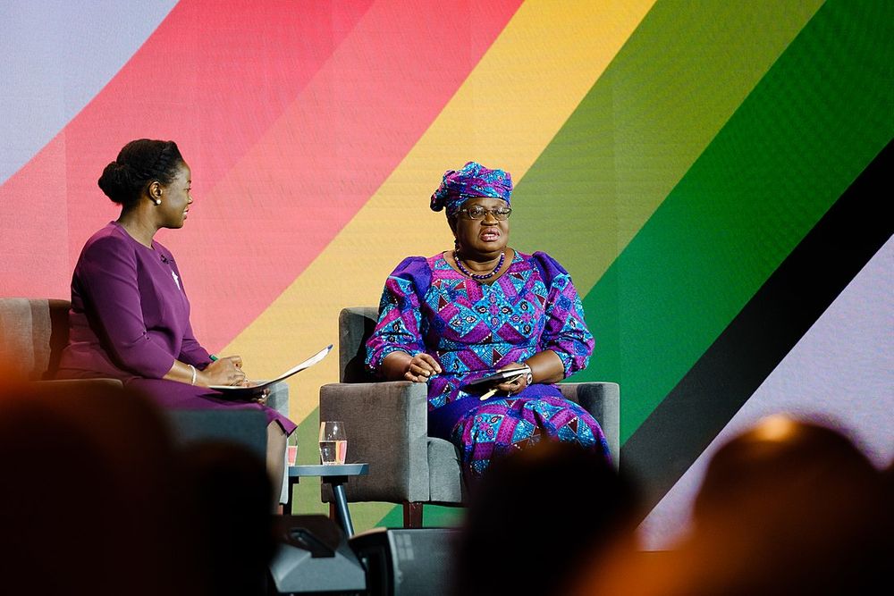 A photograph of Ngozi Okonjo-Iweala sitting on stage at the UK-Africa Investment Summit in front of a rainbow-striped background. She is wearing a colorful pink, purple, and turquoise dress and head wrap.