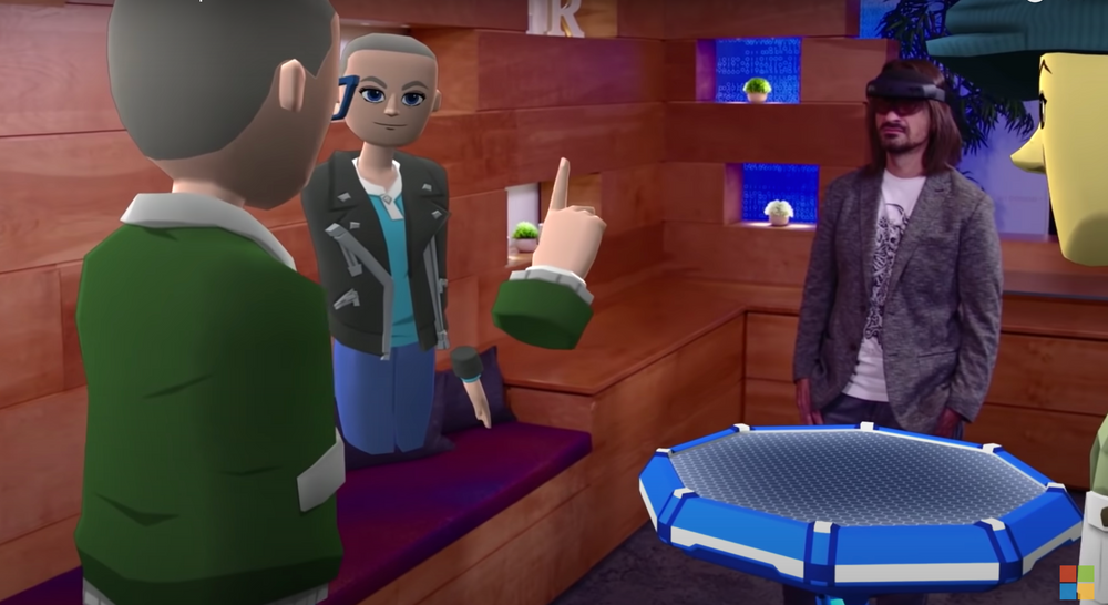 A screenshot of Microsoft Mesh's introduction video; a man is wearing a VR headset and standing in around a table talking with three cartoon avatars