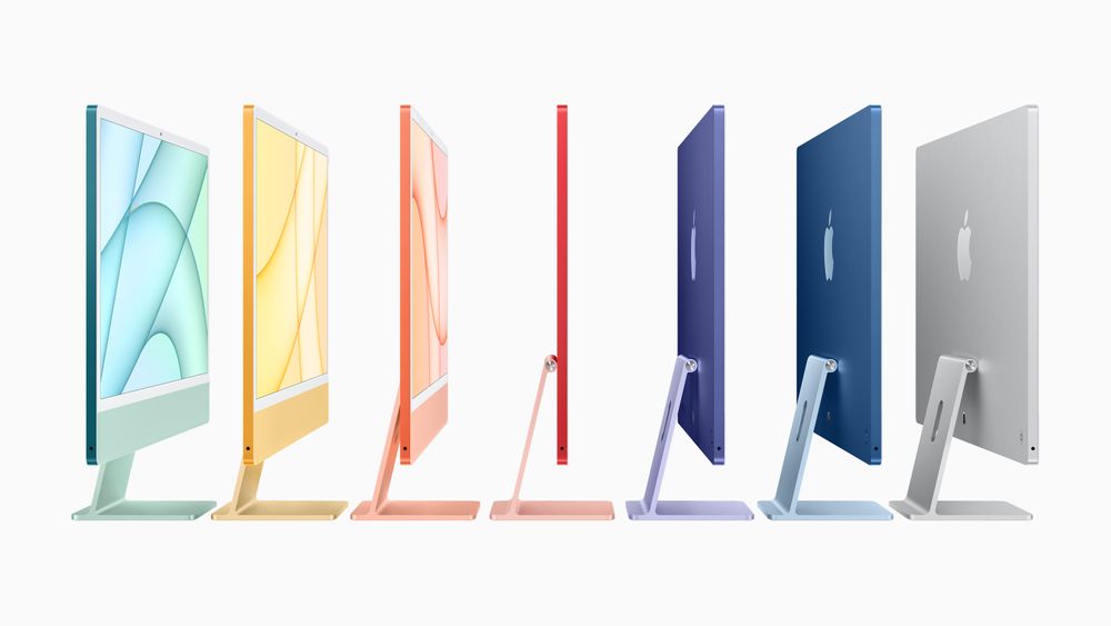 A photograph of seven Apple iMac computers in a line, photographed from the side. From left to right, the computers are turquoise, yellow, orange, red, purple, blue, and silver. 