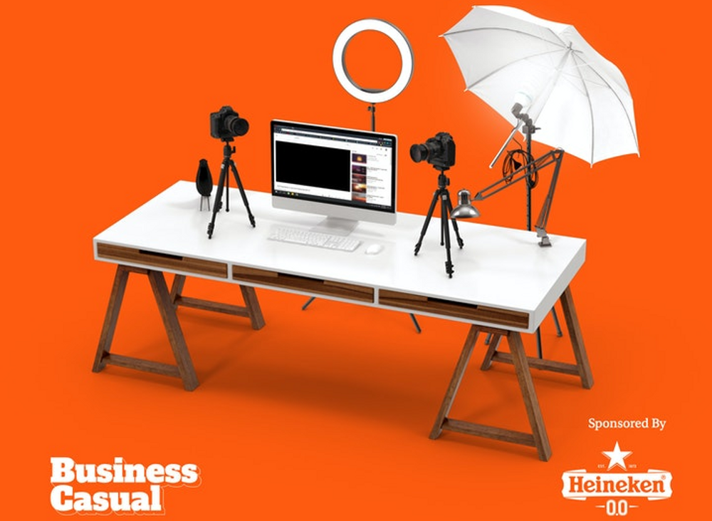 A photo of a white desk in front of an orange background. On the desk is a computer screen and keyboard, ring light, two tripods with a camera, and an umbrella reflector lighting kit.