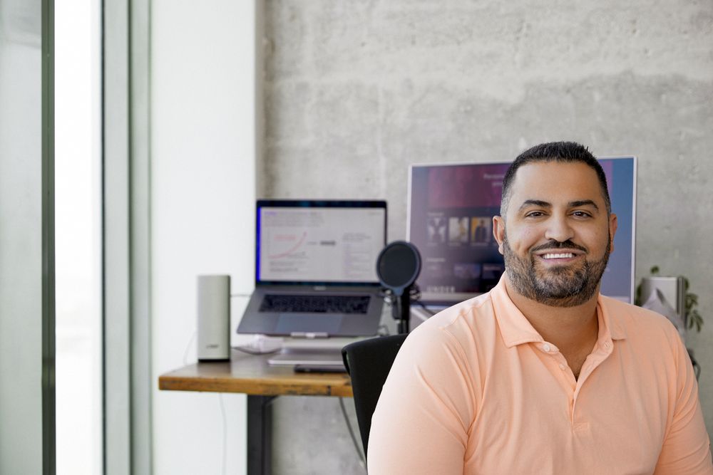 A photograph of Cameo CEO Steven Galanis. He has short cropped hair and a dark beard and is wearing a light orange t-shirt. He is sitting in front of a desk with a laptop, screen, and microphone in front of a grey stone wall.