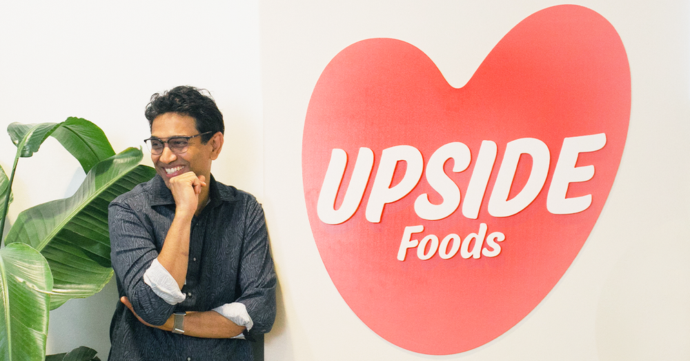 A photograph of Uma Valeti wearing a gray button down. He is smiling and resting his head on one fist. Next to him on the wall is a red blob with the name of his company, Upside Foods, written in it in white. 
