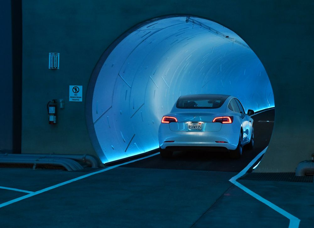 At a Boring Company debut in Las Vegas, a white Tesla sedan drives into a futuristic looking tunnel lit up blue on the inside.