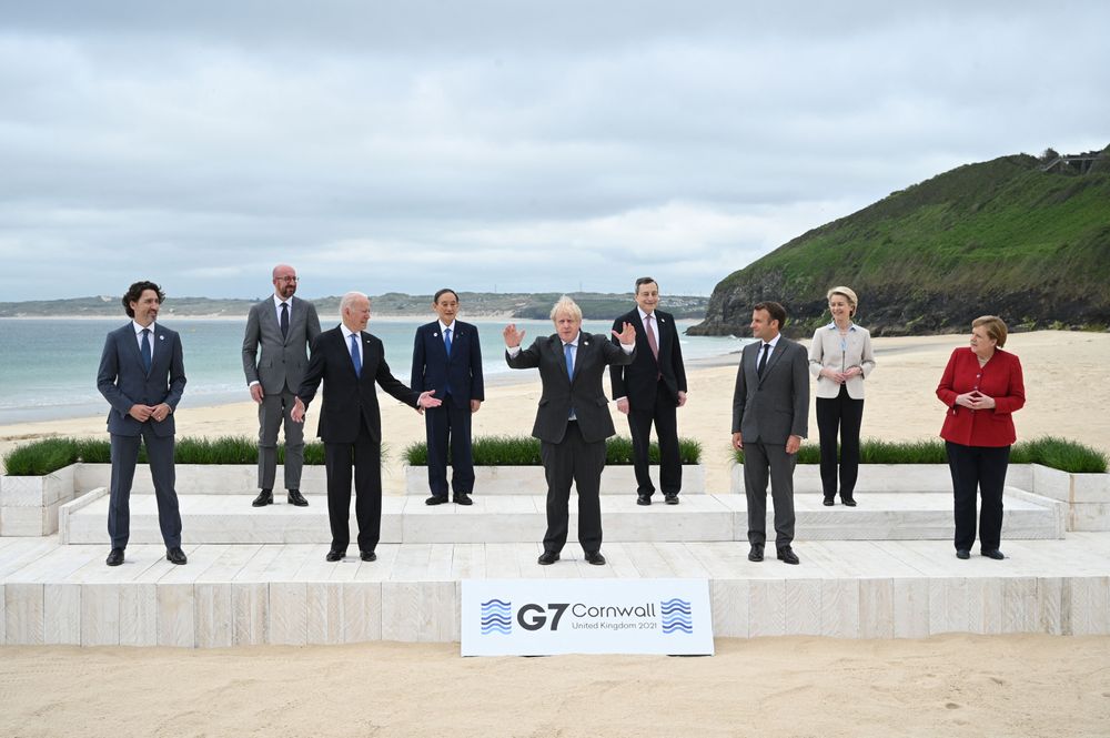 The heads of state of the G7 countries pose on a beach for a photo spaced 6' apart. In the front middle, UK PM Boris Johnson holds his hands shoulder-width apart.