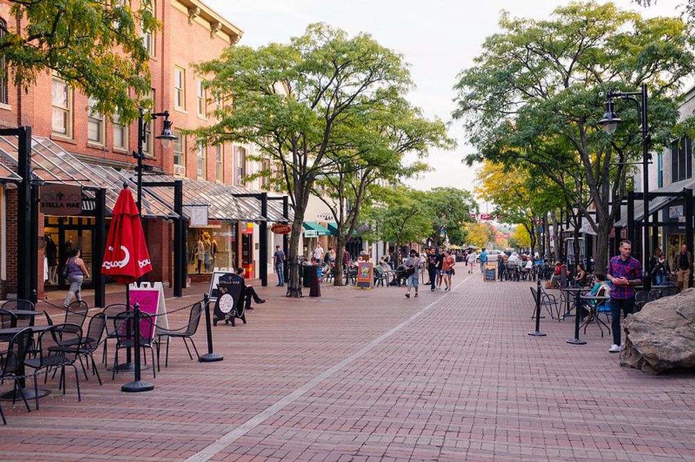 A photograph of downtown Burlington, VT. A brick-paved pedestrian walkway is filled with visitors, trees, and lined by old buildings and restaurants with patios and outdoor seating.
