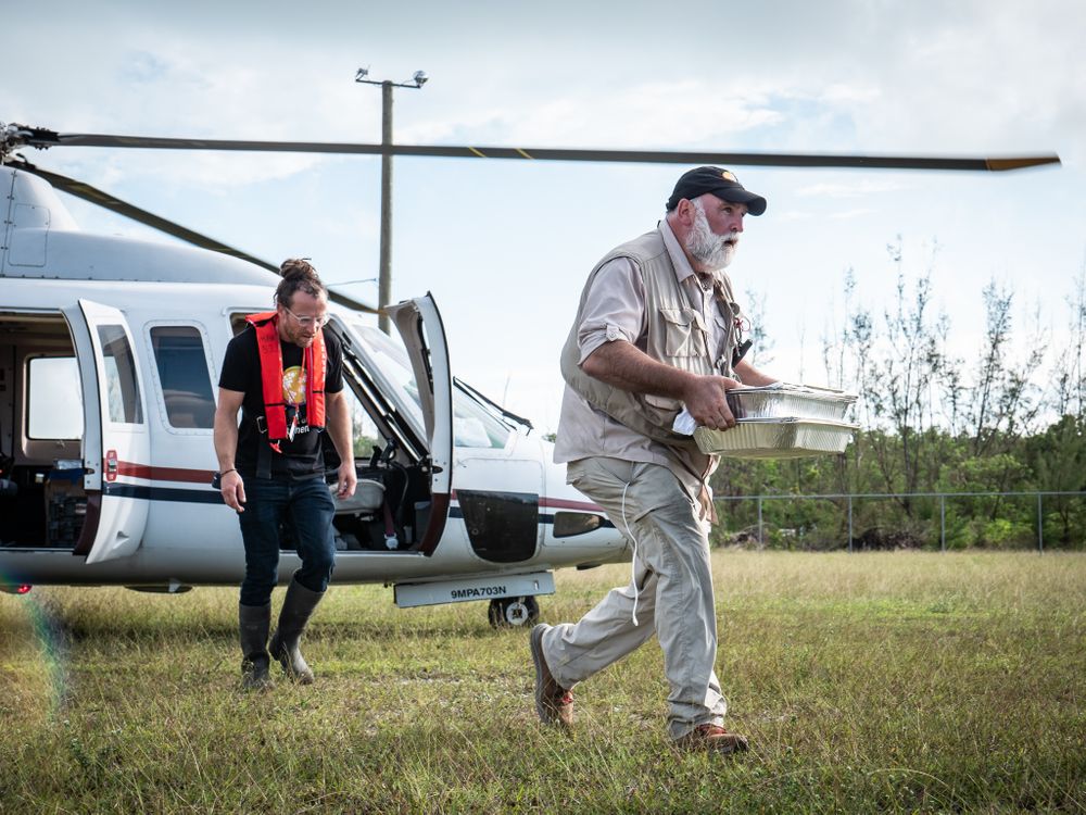 A photograph of chef Jose Andres walking away from a helicopter and carrying trays of food. He is wearing all-white and walking across a grass field. 