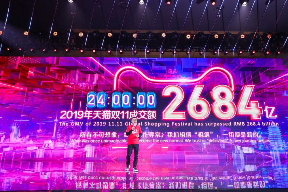 The president of Taobao speaks at Alibaba's Global Shopping Festival in front of a pink, purple, and blue screen displaying sales from Singles Day in 2019. 