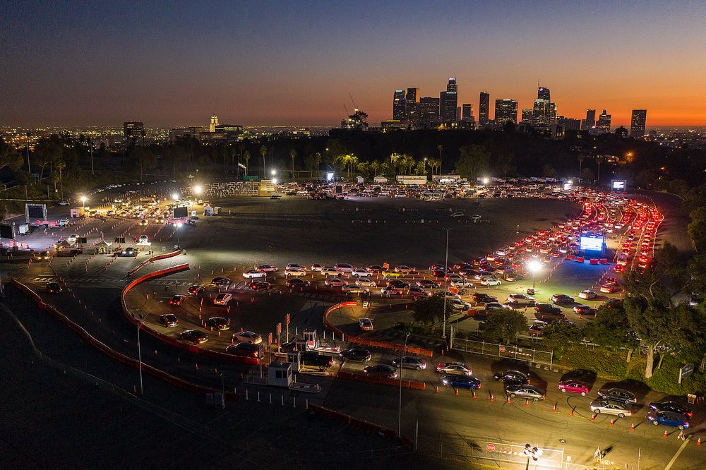 Lines of cars wrap around LA's Dodger Stadium parking lot for Covid-19 testing. The LA skyline and sunset are visible in the background.