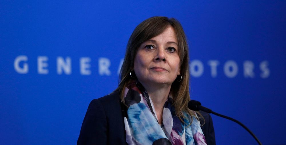 A photograph of GM CEO Mary Barra speaking into a microphone in front of a blue background. 