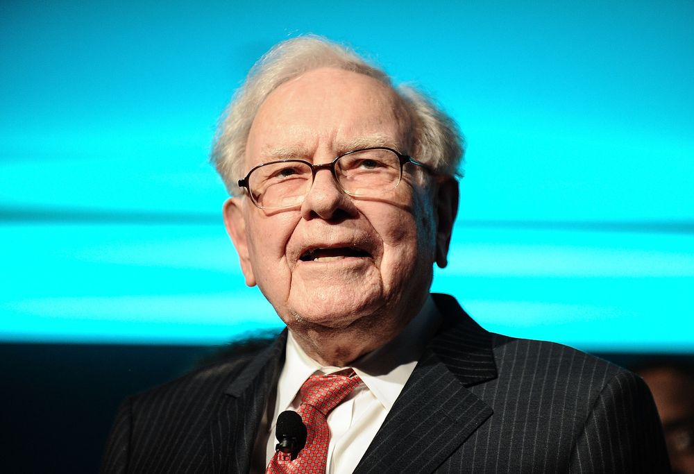 A photo of Warren Buffett from the shoulders up; He is standing in front of a turquoise background wearing a black pinstripe suit, red tie, and glasses. 