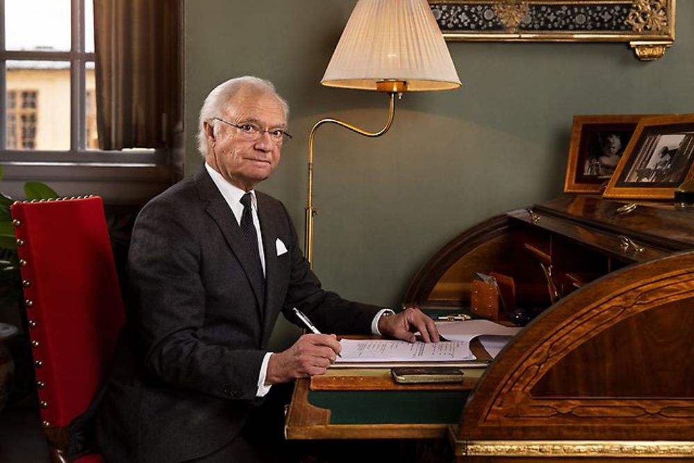 A photograph of King Carl XVI Gustaf seated at a desk signing papers. He is wearing a black suit with a tie and glasses.