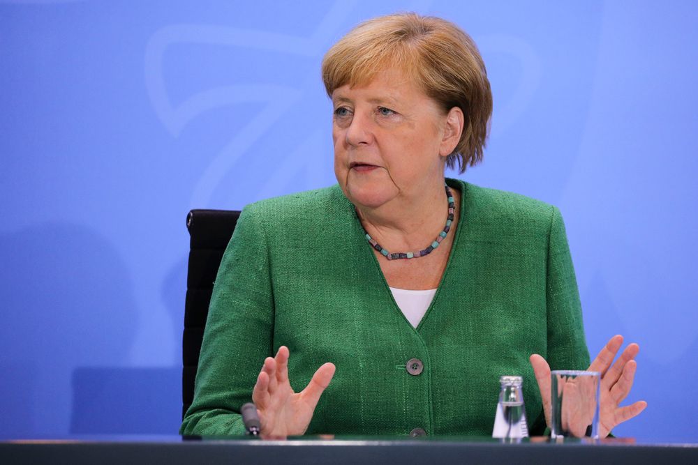 German Chancellor Angela Merkel sitting in front of a purple background during a media event. She is wearing a green suit jacket and colorful necklace. 
